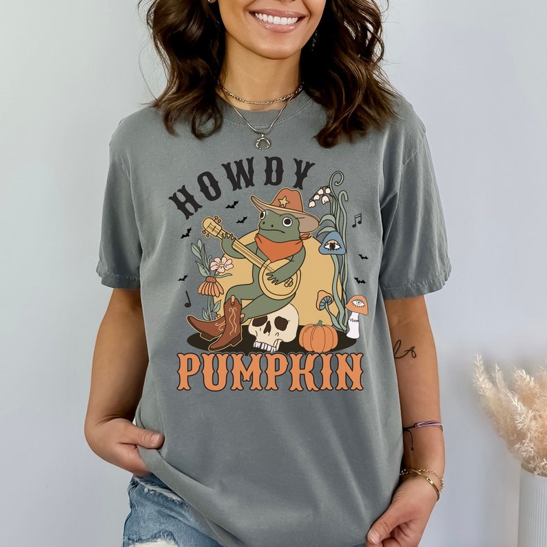 Pu&ograve; includere: Una t-shirt grigio antracite con una stampa grafica di una rana verde che indossa un cappello da cowboy e una bandana, mentre suona il banjo seduta su un teschio. Il disegno include una zucca, funghi, fiori, pipistrelli e il testo HOWDY PUMPKIN.