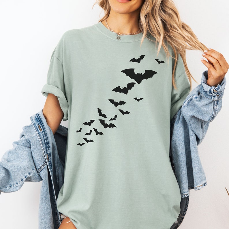 Puede incluir: Una camiseta verde claro con una silueta negra de una bandada de murci&eacute;lagos volando en diagonal por el frente.
