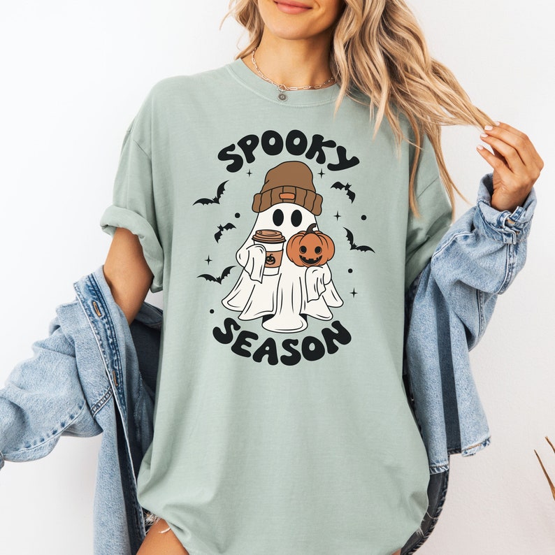 Op de afbeelding: Een lichtgroen T-shirt met een afbeelding van een geest die een beanie draagt en een pompoen en een kopje koffie vasthoudt. De tekst "Spooky Season" is onder de geest gedrukt.