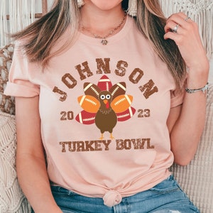 Op de afbeelding: Een roze T-shirt met een verweerde bruine afbeelding van een kalkoen met voetballen als veren. De tekst "JOHNSON 20 23 TURKEY BOWL" staat op het shirt gedrukt.