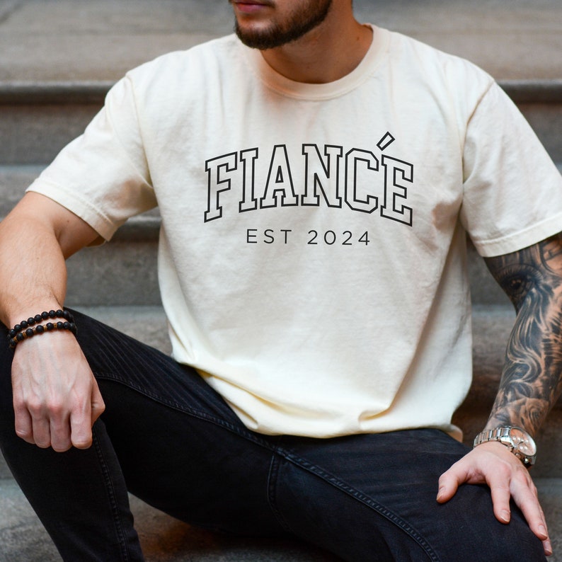 Puede incluir: Una camiseta de color crema con texto negro que dice "FIANC&Eacute; EST 2024".