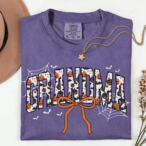 May include: A purple Comfort Colors t-shirt with the word "GRANDMA" in a Halloween-themed design. The letters are filled with pumpkins and bats, with an orange bow at the bottom. A gold star necklace and a brown hat are also in the image.