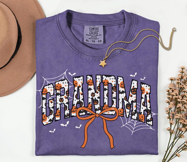 May include: A purple Comfort Colors t-shirt with the word "GRANDMA" in a Halloween-themed design. The letters are filled with pumpkins and bats, with an orange bow at the bottom. A gold star necklace and a brown hat are also in the image.