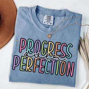 May include: A blue Comfort Colors t-shirt with the words "PROGRESS OVER PERFECTION" in colorful bubble letters. A gold necklace with a pendant is draped over the shirt. The shirt is folded and placed on a white surface.
