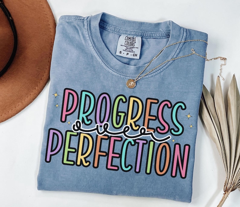 May include: A blue Comfort Colors t-shirt with the words "PROGRESS OVER PERFECTION" in colorful bubble letters. A gold necklace with a pendant is draped over the shirt. The shirt is folded and placed on a white surface.