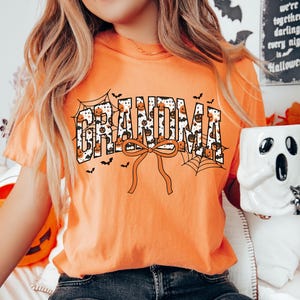 May include: Orange t-shirt with the word "GRANDMA" in a Halloween-themed design. The letters are filled with pumpkins and spiderwebs. A large orange bow is below the word, and small bats are scattered around the design. A Halloween-themed mug is held in the hand.