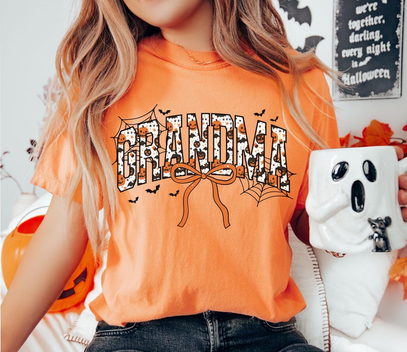 May include: Orange t-shirt with the word "GRANDMA" in a Halloween-themed design. The letters are filled with pumpkins and spiderwebs. A large orange bow is below the word, and small bats are scattered around the design. A Halloween-themed mug is held in the hand.