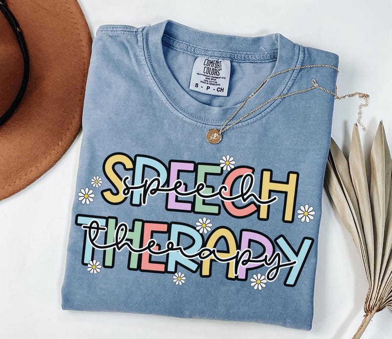 May include: A blue Comfort Colors t-shirt with the words "SPEECH THERAPY" in colorful block letters and daisy accents. A gold necklace with a pendant is draped across the shirt.