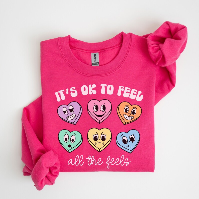 Pu&ograve; includere: Felpa rosa con il testo "It's ok to feel all the feels" e una fila di sei cuori colorati con diverse espressioni facciali.