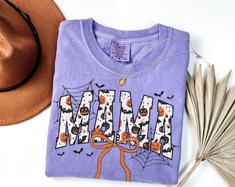 May include: A lavender Comfort Colors t-shirt with a Halloween-themed design. The word "MAMA" is printed in white with orange pumpkin and bat patterns. A gold necklace and a brown hat are also visible.