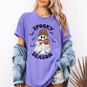 Op de afbeelding: Paars T-shirt met een cartoon-geest die een beanie draagt en een pompoen en een kopje koffie vasthoudt. De tekst "Spooky Season" staat op het T-shirt gedrukt.