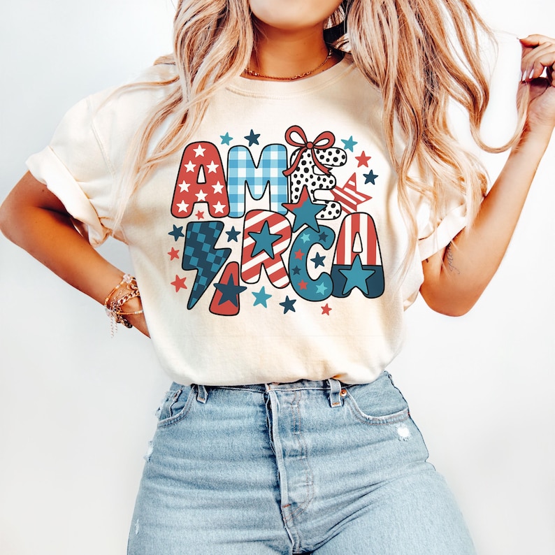 May include: Cream-coloured t-shirt featuring the word "AMERICA" in a patriotic design. The letters are decorated with red, white, and blue patterns, including stars, stripes, and gingham. A red bow and stars surround the word.