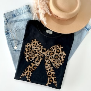 K&ouml;nnte beinhalten: Schwarzes T-Shirt mit einer gro&szlig;en Schleife im Leopardenmuster in Braun und Schwarz. Das Shirt hat einen Rundhalsausschnitt und kurze &Auml;rmel.