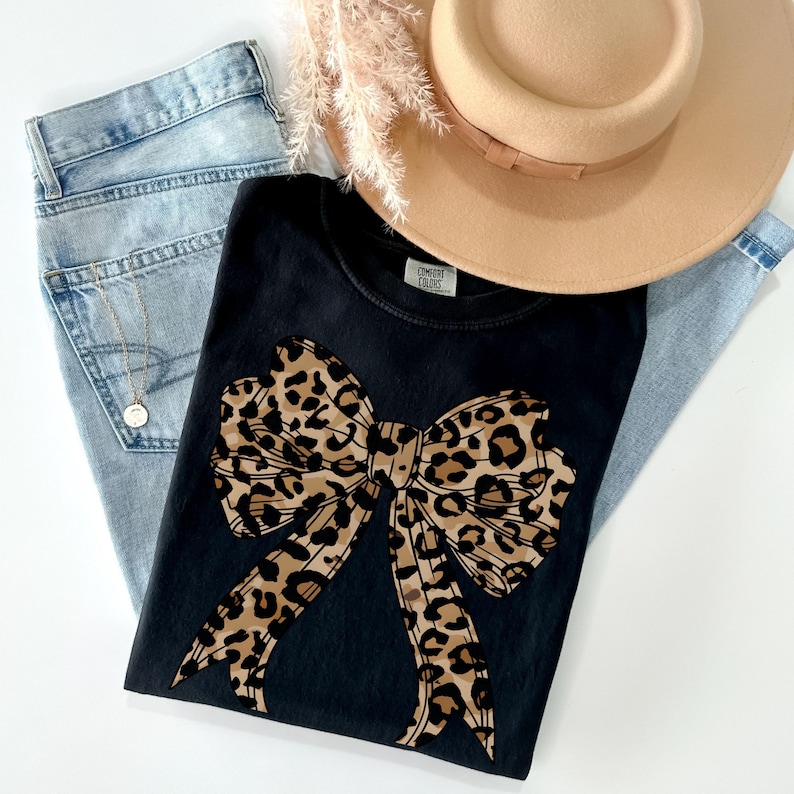 K&ouml;nnte beinhalten: Schwarzes T-Shirt mit einer gro&szlig;en Schleife im Leopardenmuster in Braun und Schwarz. Das Shirt hat einen Rundhalsausschnitt und kurze &Auml;rmel.