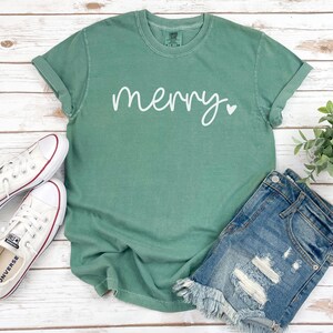 May include: A green t-shirt with the word "merry" written in white cursive with a small white heart next to it. The shirt is laying on a white wooden surface with a pair of white sneakers and a pair of denim shorts.