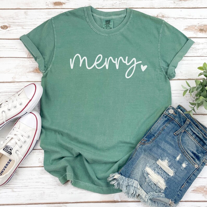 May include: A green t-shirt with the word "merry" written in white cursive with a small white heart next to it. The shirt is laying on a white wooden surface with a pair of white sneakers and a pair of denim shorts.