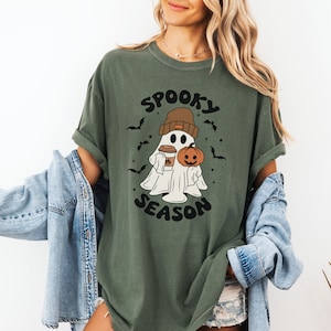 Op de afbeelding: Een groen T-shirt met een witte geest die een bruine hoed draagt en een pompoen en een kopje koffie vasthoudt. De tekst "Spooky Season" staat op het T-shirt gedrukt.