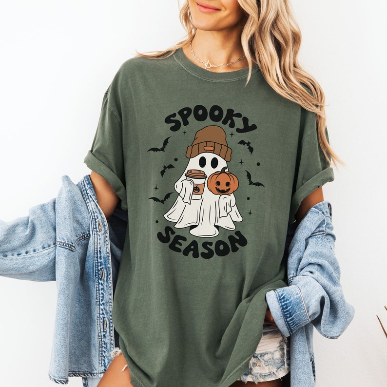 Op de afbeelding: Een groen T-shirt met een witte geest die een bruine hoed draagt en een pompoen en een kopje koffie vasthoudt. De tekst "Spooky Season" staat op het T-shirt gedrukt.