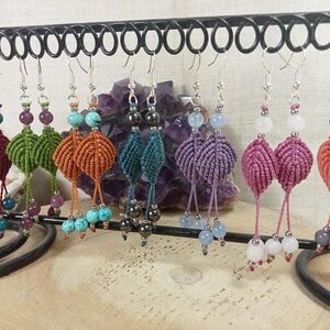 Peut inclure: Une présentation de boucles d'oreilles macramé faites à la main de différentes couleurs, dont bordeaux, vert, orange, sarcelle, violet, rose et corail. Chaque paire présente un design unique avec des accents de perles et des crochets d'oreilles argentés. Les boucles d'oreilles sont exposées sur un présentoir en métal noir.
