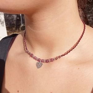 Peut inclure: Un collier bordeaux avec un pendentif en forme de cœur et des perles roses. Le collier a un cordon tressé et un charm en forme de cœur argenté avec un design complexe. Les perles roses sont entrecoupées de perles plus petites et transparentes.
