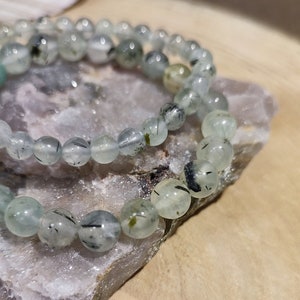 May include: Two green and black beaded bracelets with a light green and black colour scheme. The bracelets are made of round beads and are stacked on top of each other. The bracelets are sitting on a light grey rock.