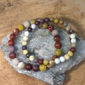 Può includere: Due braccialetti di perline con pietre dai toni della terra, tra cui rosso, marrone, giallo e crema. I braccialetti sono appoggiati su una pietra grigia, mostrando le variazioni naturali delle perline.