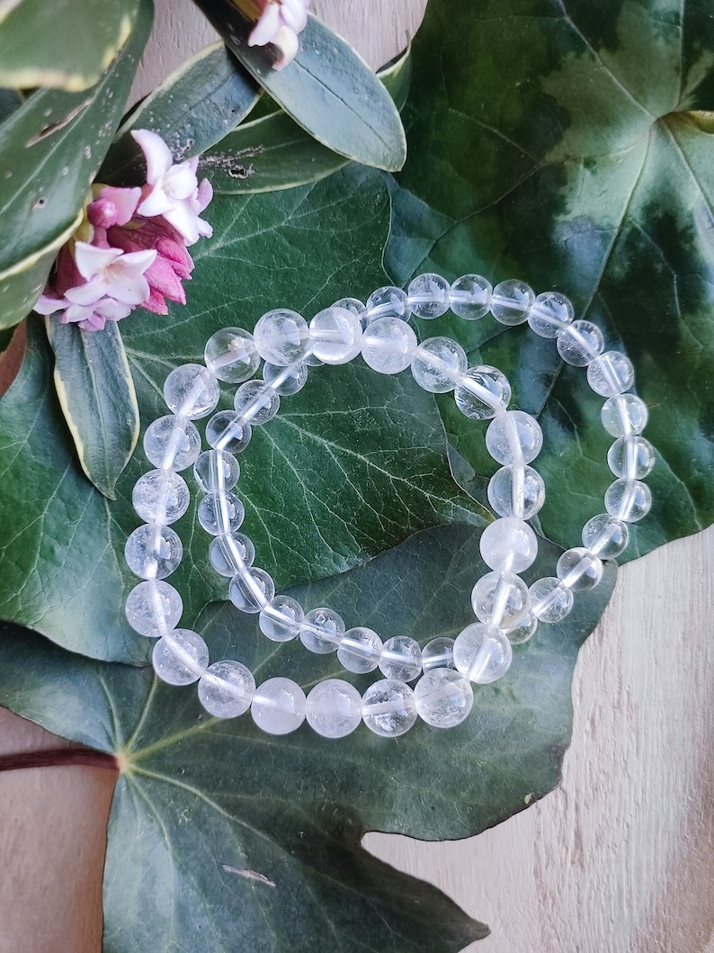 Pu&ograve; includere: Due braccialetti in cristallo di quarzo trasparente con perline rotonde, appoggiati su una foglia verde.