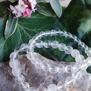 Pu&ograve; includere: Due braccialetti di perline di cristallo di quarzo trasparente su una superficie rocciosa marrone chiaro. I braccialetti sono fatti di perline rotonde e lisce.