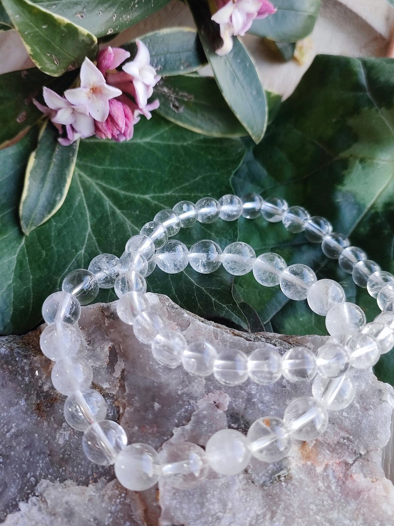 Pu&ograve; includere: Due braccialetti di perline di cristallo di quarzo trasparente su una superficie rocciosa marrone chiaro. I braccialetti sono fatti di perline rotonde e lisce.