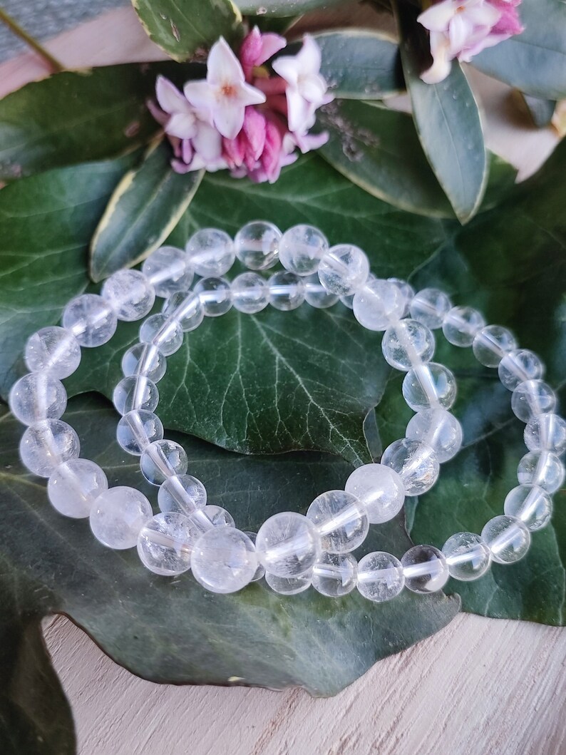 Pu&ograve; includere: Due braccialetti di perline di cristallo di quarzo trasparente su uno sfondo di foglie verdi. I braccialetti sono fatti di perline rotonde e sono impilati l'uno sull'altro.