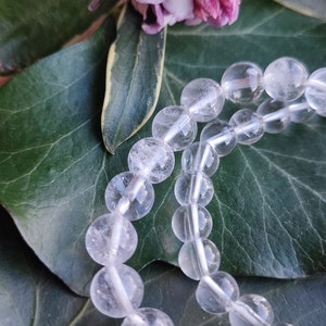 Pu&ograve; includere: Una collana di perle di cristallo di quarzo trasparente, con un aspetto naturale leggermente nuvoloso, &egrave; esposta su uno sfondo di foglie verdi.
