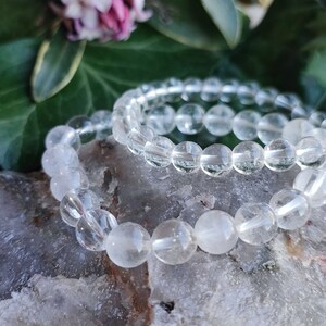 Pu&ograve; includere: Due braccialetti di cristallo di quarzo trasparente impilati uno sopra l'altro. I braccialetti sono fatti di perline rotonde e riposano su una superficie grigia testurizzata.