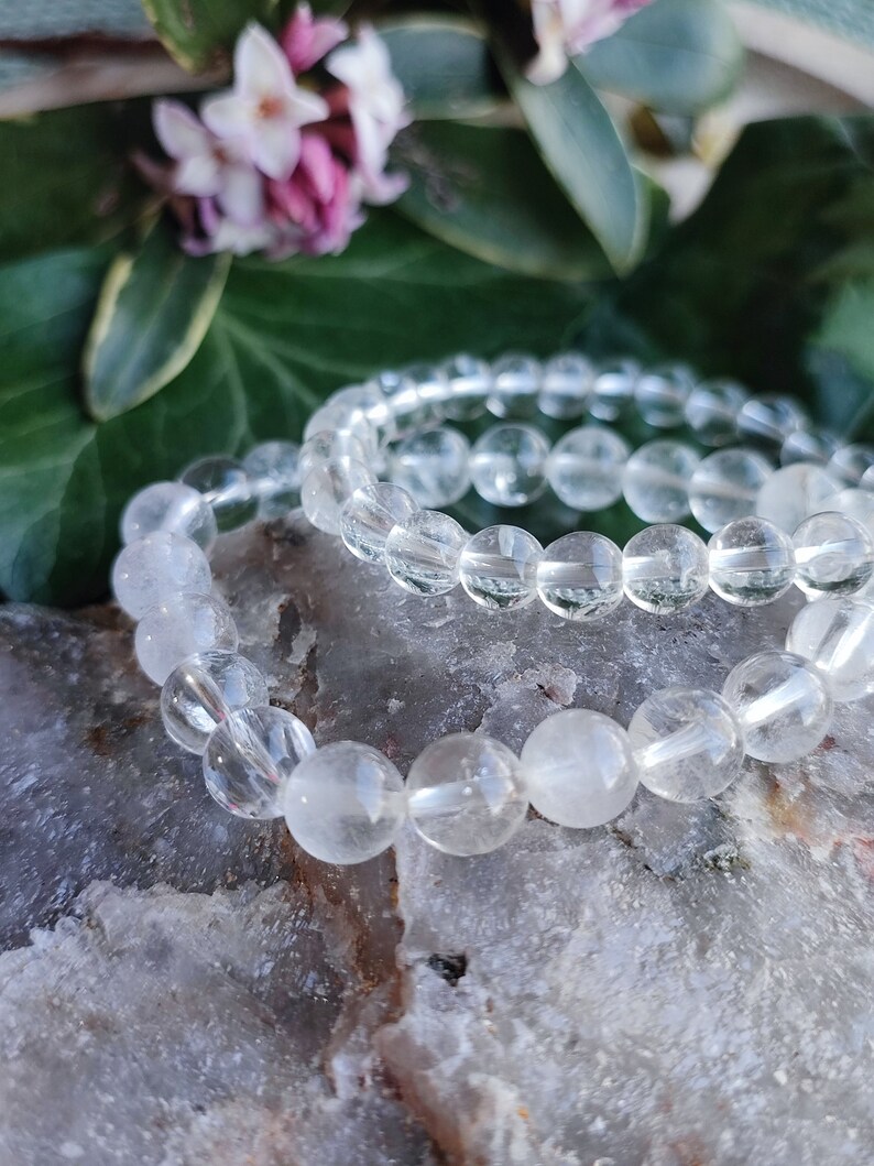 Pu&ograve; includere: Due braccialetti di cristallo di quarzo trasparente impilati uno sopra l'altro. I braccialetti sono fatti di perline rotonde e riposano su una superficie grigia testurizzata.
