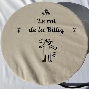 以下が含まれることがあります： 帽子をかぶった棒人間と「Le roi de la billig」という文字が書かれた、ベージュの丸いクッション。クッションには黒いコードが付いている。