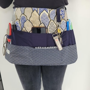 Peut inclure: Une ceinture utilitaire en tissu à motif bleu et or avec plusieurs poches et un porte-clés. La ceinture a une poche en tissu bleu avec un carnet et un stylo, une poche pour téléphone et une poche pour clés. La ceinture est portée par une personne.