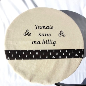 Peut inclure: Une housse ronde en tissu beige avec une bordure de ruban noir et le texte "Jamais sans ma billig" en lettres noires. Le ruban a un motif répété de triskèles blancs sur fond noir.
