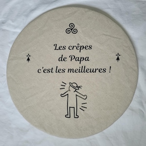 以下が含まれることがあります： 「Les crêpes de Papa c'est les meilleures!」の黒い文字と棒人間のイラストが描かれた、丸いベージュ色の布製品。ケルトノットと2つの小さな装飾シンボルも特徴です。