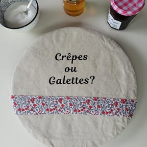 Peut inclure: Une housse ronde en tissu beige avec une bordure florale rouge et blanche et le texte "Crêpes ou Galettes?" brodé en noir.