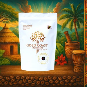 May include: A white bag of Gold Coast Roasters coffee beans from Ghana. The bag features a hand-drawn illustration of a coffee tree and a bee. The bag is sitting on a wooden surface with coffee beans scattered around it.