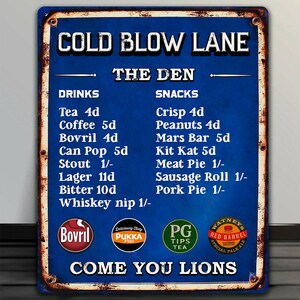 Pode incluir: Uma placa de metal azul vintage com o texto "COLD BLOW LANE" e "THE DEN". Lista bebidas e snacks com preços, incluindo chá e uma barra Mars. A placa tem uma borda enferrujada e desgastada e a frase "COME YOU LIONS".