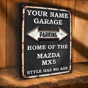 May include: A rectangular metal sign with a black background and white text. The sign reads "YOUR NAME GARAGE" above an arrow pointing left and right with the word "PARKING" in the center. Below the arrow, it says "HOME OF THE MAZDA MX5" and "STYLE HAS NO AGE".