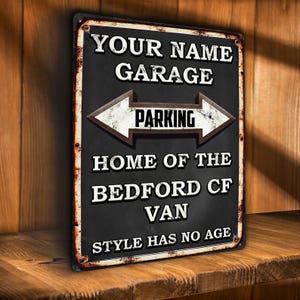 May include: A rectangular black metal sign with white text that reads "YOUR NAME GARAGE PARKING HOME OF THE BEDFORD CF VAN STYLE HAS NO AGE". The sign has a distressed, vintage look with a brown arrow.