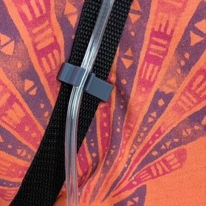 May include: A close-up of a black strap with a clear plastic tube running through it. The strap is attached to a piece of clothing with a geometric pattern in shades of orange, red, and purple.