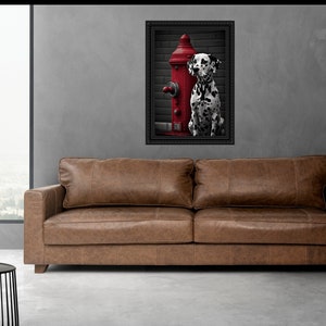 May include: A black and white Dalmatian dog sits next to a red fire hydrant in a framed photograph. The photograph is hanging on a grey wall above a brown leather sofa.