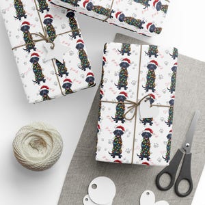 May include: White wrapping paper featuring a repeating pattern of black Labrador dogs wearing red Santa hats and wrapped in colorful Christmas lights, accented with grey paw prints and pink bone shapes, tied with twine and white star-shaped gift tags.