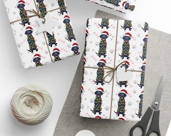 Pode incluir: Papel de embrulho branco com um padrão repetitivo de cães Labrador pretos usando gorros de Pai Natal vermelhos e enrolados em luzes de Natal coloridas, acentuado com pegadas cinzentas e formas de ossos cor-de-rosa, atado com cordel.