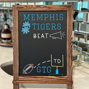 May include: A brown wooden chalkboard easel with a black chalkboard surface. The chalkboard has white text that reads "Memphis Tigers Beat" with a drawing of a megaphone. Below the text is a drawing of a football being thrown towards a goal post with the text "GTG TD".