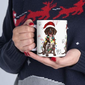 May include: White ceramic mug featuring a brown and white dog wearing a Santa hat and Christmas lights. The mug has paw prints and a bone design. A festive holiday-themed gift.