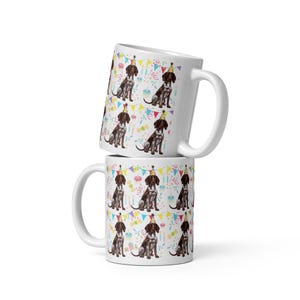 May include: White ceramic mug featuring a colourful pattern of German Shorthaired Pointers wearing party hats and surrounded by birthday decorations. The mug is stacked on top of another identical mug.