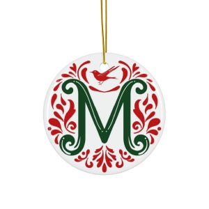 May include: A white ceramic Christmas ornament with a gold string. The ornament features a large green letter "M" in the center, surrounded by red floral designs and a red bird. The ornament is round.